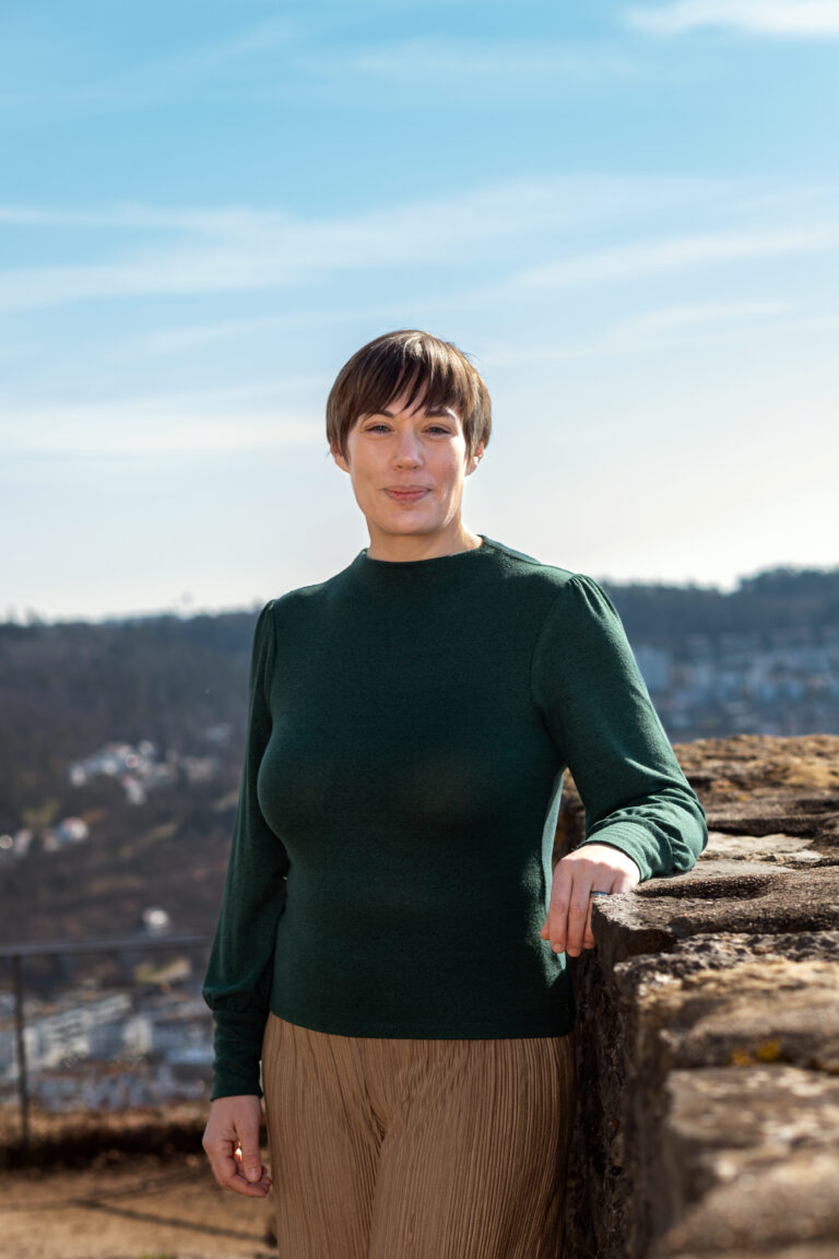 Dorothee Hildbrand steht gelassen im Sonnenlicht, angelehnt an eine Mauer – ein Moment von Ruhe, Klarheit und innerer Stärke.