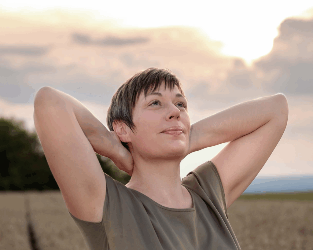 Dorothee Hildbrand steht entspannt auf einem Feld, legt die Hände hinter den Kopf und blickt ruhig in den Himmel – Symbol dafür, Grübeln zu stoppen und innere Ruhe zu finden