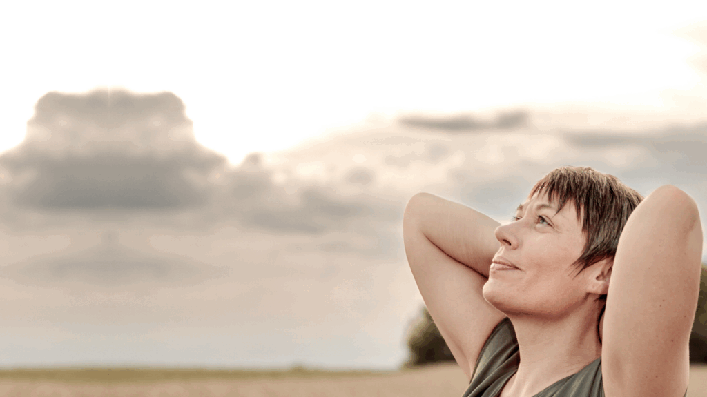 Dorothee Hildbrand steht entspannt auf einem Feld, legt die Hände hinter den Kopf und blickt ruhig in den Himmel – Symbol dafür, Grübeln zu stoppen und innere Ruhe zu finden