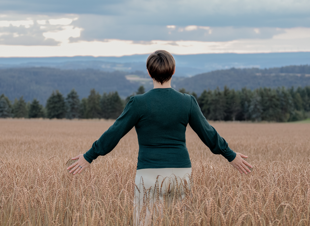 Dorothee Hildbrand steht mit ausgebreiteten Armen in einem Getreidefeld und blickt in die Weite – Symbol für ihren Wendepunkt vom Grübeln zur inneren Ruhe.