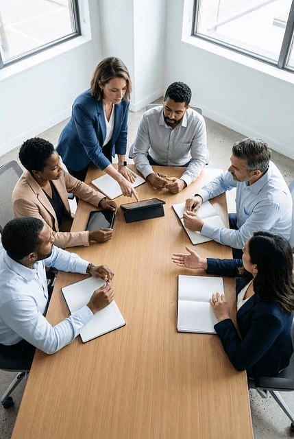Diverse team of six professionals collaborating around a conference table with notebooks and a tablet in a bright room.